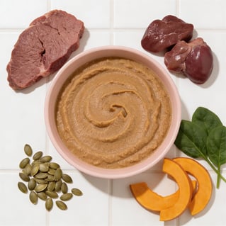 Bowl of Wundercat Beef Pâté surrounded by fresh ingredients including beef, beef liver, beef heart, pumpkin, spinach, and pumpkin seeds on a white tiled surface.
